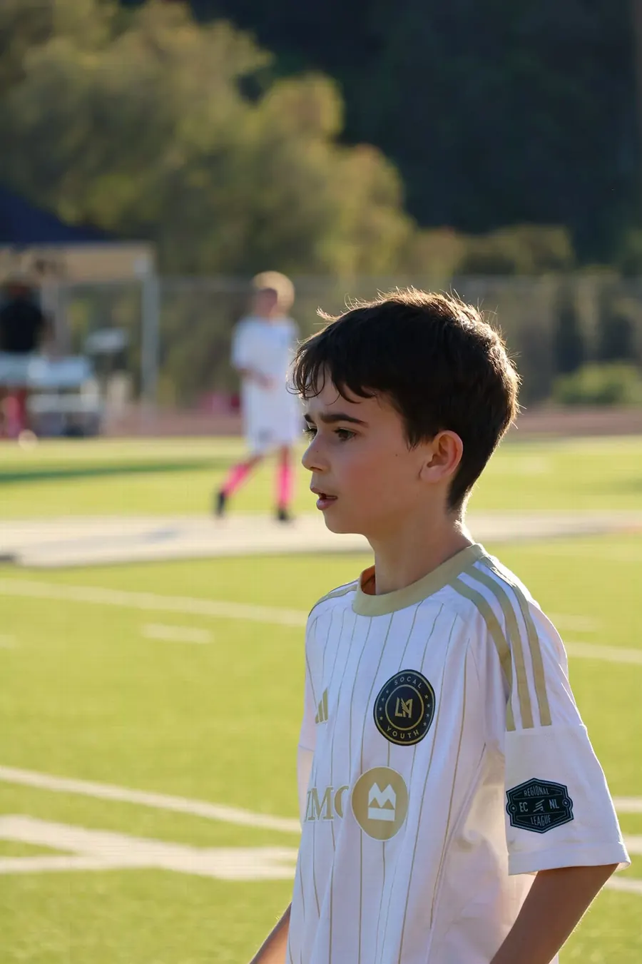 Young soccer player in profile on a sunlit field with a teammate blurred behind, photographed by Lihi Matzri